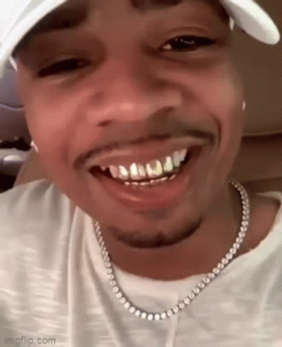 A Close Up Of A Man Wearing A Hat And A Necklace With His Teeth Visible . GIF