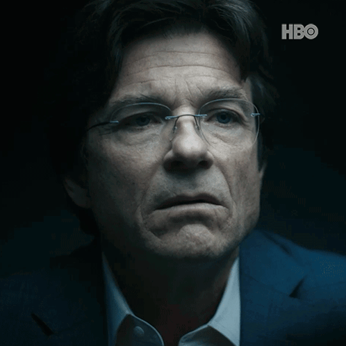 A Close Up Of A Man Wearing Glasses And A Suit With The Hbo Logo Behind Him GIF