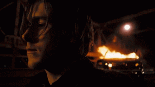 A Close Up Of A Person 'S Face With A Burning Vehicle In The Background GIF