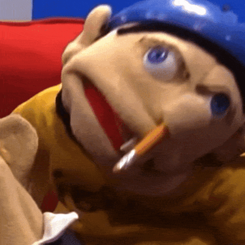 A Close Up Of A Puppet Wearing A Yellow Shirt And A Blue Helmet With A Cigarette In His Mouth GIF