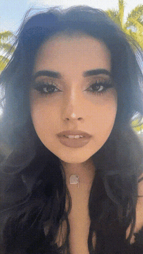 A Close Up Of A Woman 'S Face With A Necklace Around Her Neck And A Palm Tree In The Background GIF