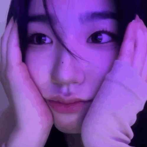 A Close Up Of A Woman 'S Face With Her Hands On Her Face In Front Of A Purple Light . GIF