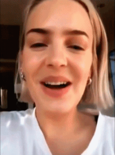 A Close Up Of A Woman 'S Face With Short Blonde Hair And A White Shirt Smiling . GIF