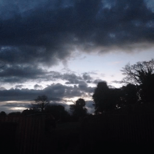 A Cloudy Sky With Trees In The Foreground And A Fence In The Background GIF