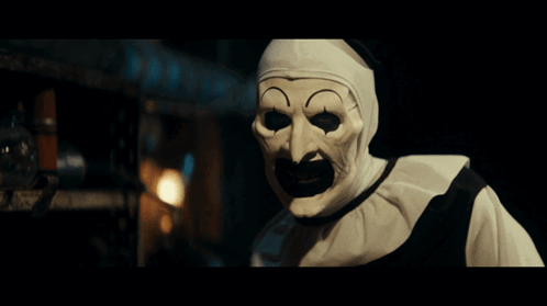 A Clown Wearing A White Mask With A Black Mouth Is Standing In A Dark Room GIF