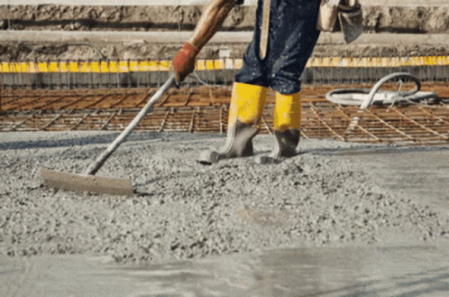A Construction Worker Wearing Yellow Boots Spreads Concrete With A Rake GIF