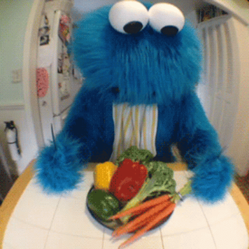 A Cookie Monster Is Sitting At A Table With A Plate Of Vegetables In His Mouth GIF