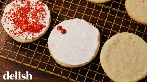 A Cookie With White Frosting And Red Sprinkles Sits On A Wire Rack With The Word Delish Below It GIF
