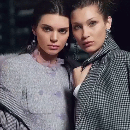 A Couple Of Women Standing Next To Each Other Wearing Coats And Earrings . GIF