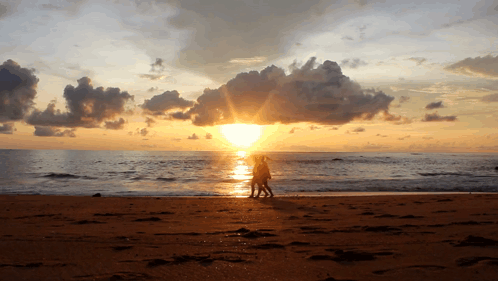 A Couple Walking On A Beach At Sunset With The Sun Shining Through The Clouds GIF