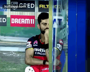 A Cricket Player Is Sitting In Front Of A Glass Door With His Hand On His Chin . GIF