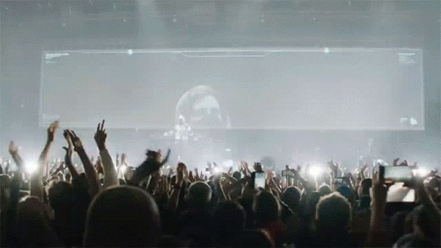 A Crowd Of People At A Concert With Their Hands In The Air In Front Of A Large Screen With A Skull On It GIF