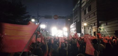 A Crowd Of People Holding Up Flags In Front Of A Building That Says ' Sydney Bank ' On It GIF