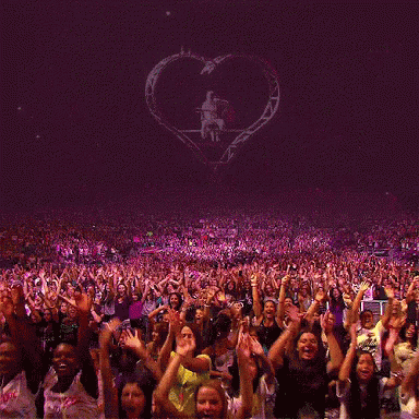 A Crowd Of People With Their Hands In The Air At A Concert With A Heart In The Background GIF