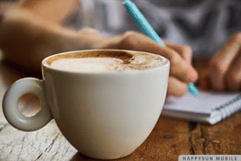 A Cup Of Coffee Sits On A Table Next To A Person Writing On A Notebook GIF
