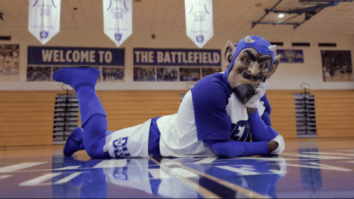 A Devil Mascot Is Laying On The Floor In Front Of A Sign That Says Welcome To The Battlefield GIF