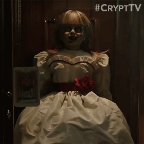 A Doll In A White Dress Is Sitting In A Chair Holding A Picture Of Annabelle . GIF