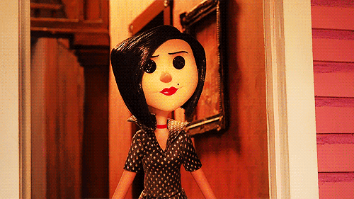 A Doll With Black Hair And Red Lips Stands In Front Of A Pink House GIF