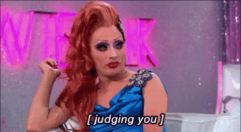 A Drag Queen In A Blue Dress Is Sitting In Front Of A Pink Sign And Says `` Judging You '' . GIF