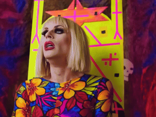 A Drag Queen In A Colorful Dress Stands In Front Of A Neon Sign With A Cross On It GIF