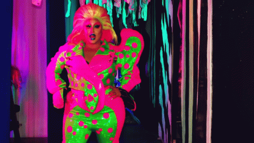 A Drag Queen In A Neon Pink And Green Outfit Is Standing In A Dark Hallway . GIF