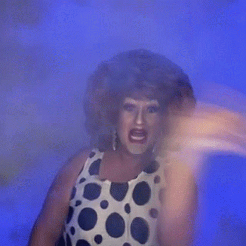 A Drag Queen In A Polka Dot Dress Is Standing In Front Of A Blue Background And Making A Funny Face . GIF