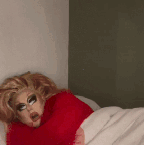 A Drag Queen In A Red Sweater Is Laying In Bed GIF