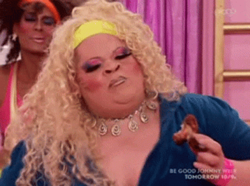 A Drag Queen Is Eating A Piece Of Chicken While A Woman Looks On . GIF