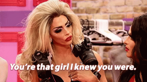 A Drag Queen Is Holding A Camera In Her Hand And Talking To Another Woman . GIF
