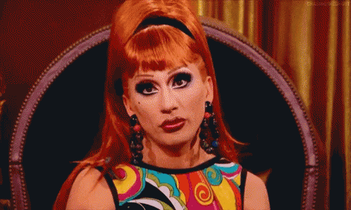 a drag queen is sitting in a chair wearing a colorful shirt and earrings .