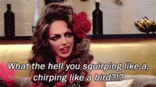 A Drag Queen Is Sitting On A Couch With A Flower In Her Hair GIF