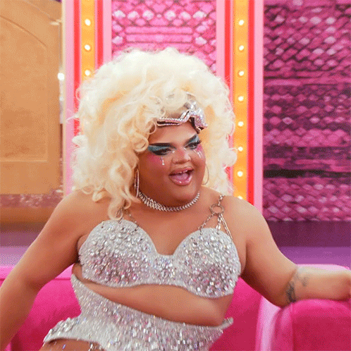 Drag Queen Sitting On Pink Couch Wearing Bra And Necklace GIF