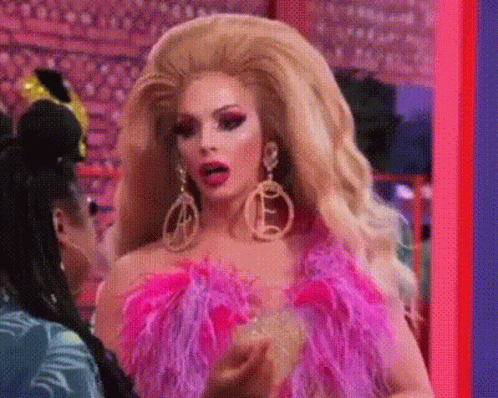 A Drag Queen Is Wearing A Pink Feathered Top And Earrings And Talking To Another Drag Queen . GIF