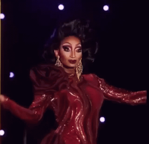 A Drag Queen Is Wearing A Red Dress And Earrings While Standing On A Stage . GIF