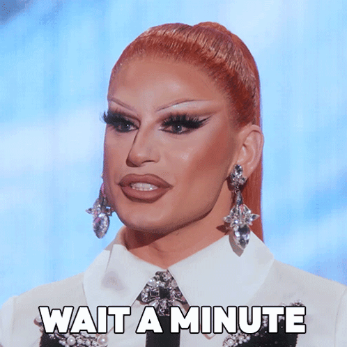 A Drag Queen Says Wait A Minute While Wearing Earrings And A White Shirt GIF