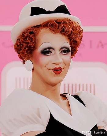 A Drag Queen Wearing A White Hat And Earrings GIF