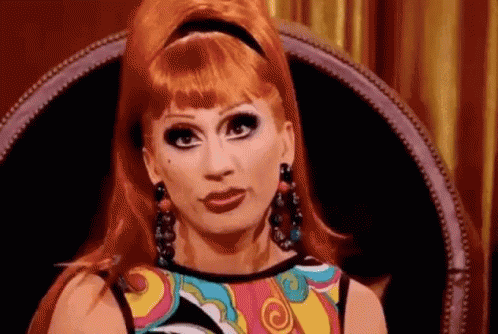 a drag queen with red hair is sitting in a chair and making a funny face .