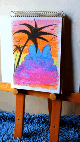 A Drawing Of A Sunset With Palm Trees Is On A Wooden Easel GIF