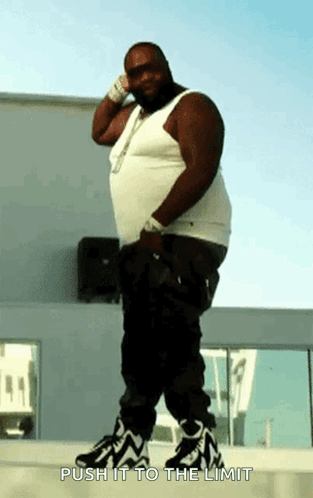 A Fat Man In A White Tank Top And Black Pants Is Standing On A Sidewalk With The Words Push It To The Limit Above Him GIF
