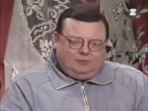 A Fat Man Wearing Glasses And A Blue Jacket Is Sitting In Front Of A Window . GIF