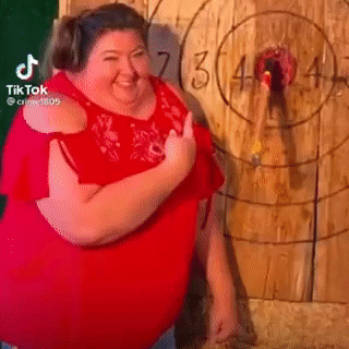 A Fat Woman In A Red Shirt Is Standing In Front Of A Wooden Target . GIF