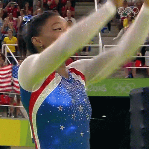 A Female Gymnast In A Blue Red And White Leotard With The Olympics Logo In The Background GIF