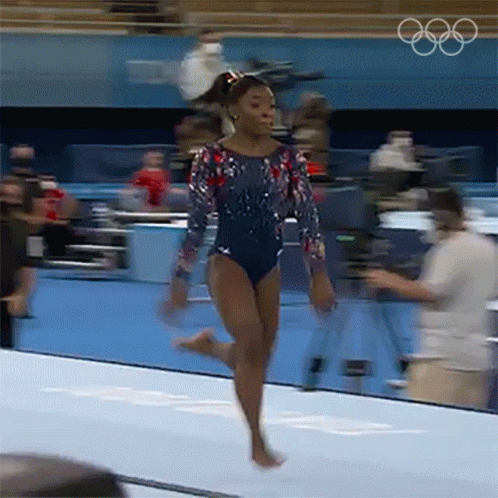 A Female Gymnast Is Doing A Trick On A Mat With The Olympic Rings In The Background . GIF