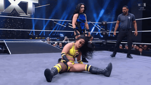 A Female Wrestler Is Laying On The Ground In A Wrestling Ring While Another Wrestler Stands Behind Her GIF