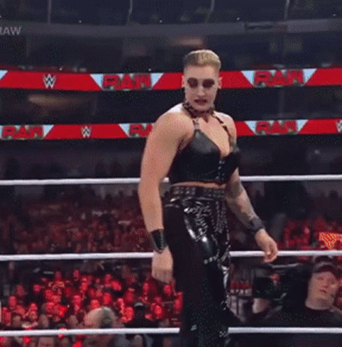 A Female Wrestler Stands In A Wrestling Ring With A Crowd Watching And A Sign That Says Raw GIF