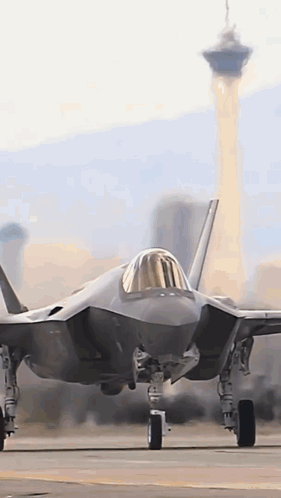 A Fighter Jet Is Sitting On A Runway With A City In The Background . GIF