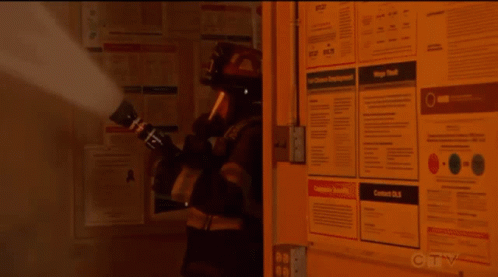 A Fireman Is Spraying Smoke In Front Of A Wall With Posters On It Including One That Says Contact Us GIF
