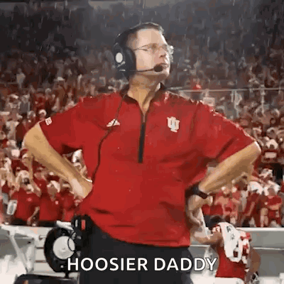 A Football Coach Wearing Headphones And A Red Shirt Is Standing On The Sidelines Of A Field . GIF