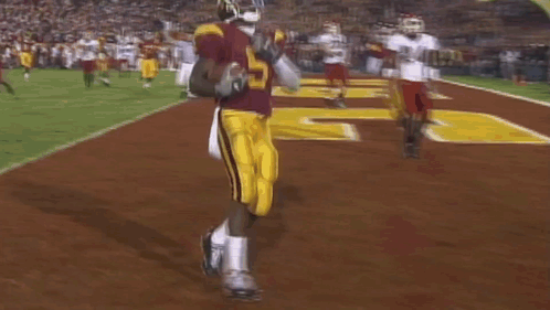 A Football Player In A Maroon Uniform Is Holding A Football In His Hand While Standing On A Field . GIF