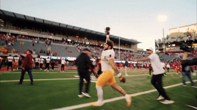 a football player is running on a field with a stadium in the background that says " trophy room "